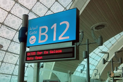 Ein Schild am Flughafen Dubai (DXB), das in Weiß auf Himmelblau das Gate B12 anzeigt. Eine digitale Anzeige darunter weist auf den Flug EK725 nach Daressalam hin.