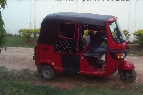 Ein rotes Bajaj (auch Bajaji oder Autorikscha) mit schwarzem Dach. Das Bajaj steht auf einem Sandweg vor einer weißen Mauer. Ein Bajaj ist ein motorisiertes Fahrzeug mit drei Rädern.