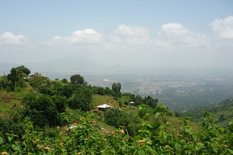 Blick vom Uluguru-Gebirge ins Tal und auf die nahe gelegene Stadt Morogoro. Im Vordergrund ist ein Hügel mit starker grüner Vegetation zu sehen.