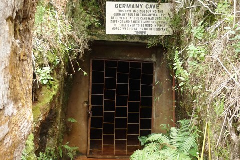 Das Foto zeigt den Eingang zu einer vergitterten Höhle mit einem Schild, auf dem steht "Germany Cave. This cave was dug during the Germany rule in Tanganyika. it is believed that the cave was used in defence and bauxite mine searching. it is believed to be used in first world war 1914-1918 by German troupe"